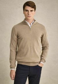 Beige Reißverschluss-Pullover aus weichem Stoff, mit hohem Kragen, gerippten Bündchen und Saum, kombiniert mit dunkelblauen Hosen.