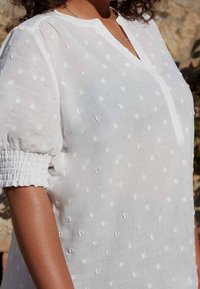 Donna che indossa una blusa bianca a pois con maniche corte a sbuffo e polsini arricciati, su uno sfondo esterno sfocato.