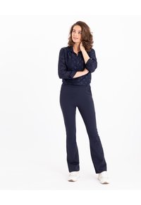 Navy blouse met gestructureerd hartpatroon en knoopsluiting, gecombineerd met strakke navy broek en witte sneakers, staande op een witte achtergrond.