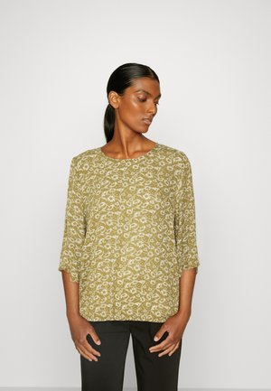 Olivengrønn bluse med hvit blomstermønster, rund hals, trekvart ermer og løs passform. Båret med svarte bukser.