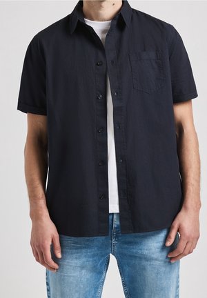 Homme portant une chemise noire à manches courtes déboutonnée par-dessus un t-shirt blanc avec un jean bleu, debout devant un fond clair uni.