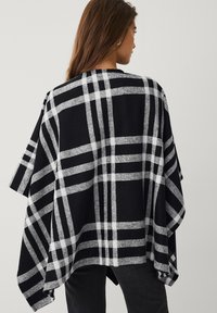 Sort og hvid ternet poncho lavet af blødt stof, med et oversized design, brede ærmer og asymmetrisk kant.