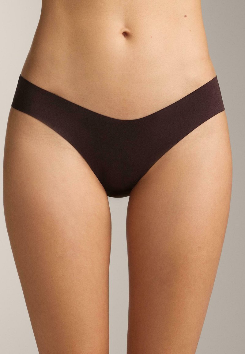 Vrouwelijke torso in naadloze donkerbruine bikini-achtige onderbroek tegen een neutrale achtergrond.
