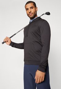Crna majica s kapuljačom od glatkog materijala, s prednjim džepom, kapuljačom s vezicom i uvezenim logotipom. Nosena s plavim golf hlačama i drži golf palicu.