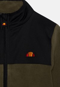 Polarowa kurtka z czarną górną częścią i oliwkowo-zielonym dolnym. Posiada zamek błyskawiczny oraz haftowane logo w kolorze czerwonym, żółtym i czarnym.