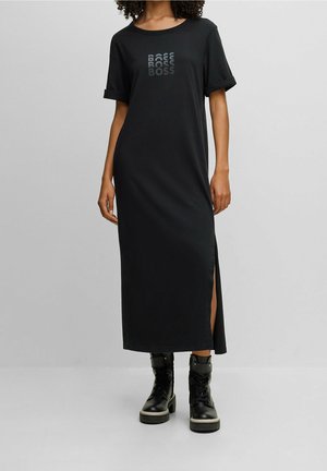Vrouw in een zwarte midi-jurk met korte mouwen en een split aan de zijkant, met het "BOSS" logo op de borst, gecombineerd met zwarte veterschoenen tot de enkel.