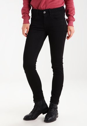 Kobieta ubrana w czarne obcisłe jeansy, czarne sznurowane botki za kostkę oraz czerwoną pikowaną bluzkę z długim rękawem, stojąca na białym tle.