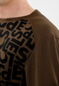 Braunes T-Shirt mit einem großen schwarzen Textgrafik. Der Stoff ist glatt, mit kontrastierenden strukturierten Akzenten entlang der Schulternaht.