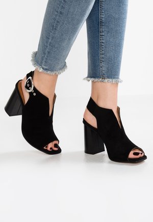 Czarne zamszowe buty na obcasie z odkrytymi palcami i wycięciami, ozdobione srebrną klamrą oraz teksturowanym, klockowym obcasem. Jeansy ze postrzępionymi mankietami.