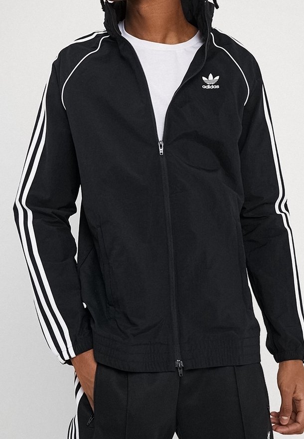 Svart zip-up-jacka av lättviktsmaterial. Har vita ränder längs ärmarna och en liten logotyp på bröstet. Justerbar huva ingår.