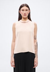 Blúzka bez rukávov z ľahkej broskyňovej látky, s okrúhlym výstrihom a hladkou textúrou. Nosená s čiernymi nohavicami, ktoré odhaľujú detaily tetovania na ruke.