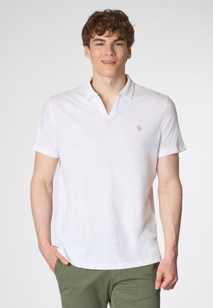 Giovane uomo con capelli ricci sorride, indossando una maglietta polo bianca e pantaloni verdi, su uno sfondo grigio chiaro.