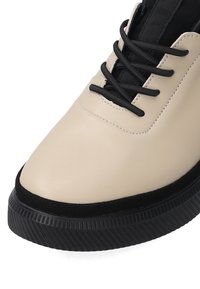 Beige Leder Sneaker mit einer strukturierten schwarzen Gummisohle. Verfügt über schwarze Schnürsenkel und eine Stoffzunge für einen sicheren Sitz. Rundes Zehen-Design.