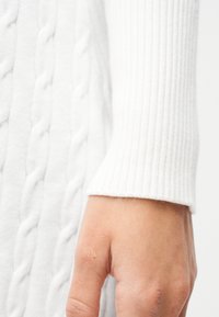 Κλείσιμο πλάνου ενός χεριού με ανοιχτόχρωμο δέρμα που φοράει μανίκι άσπρου ριγωτού πουλόβερ, σε φόντο λευκού πλεκτού υφάσματος με καλώδια.