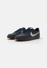 Nike sportbačiai tamsiai mėlynos odos su smėlio spalvos akcentais, turintys žemą profilį, apvalią nosį, su raišteliais ir tekstūruota ruda guma.