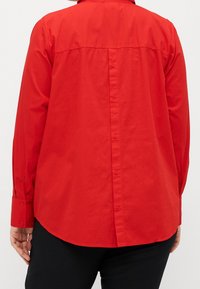 Persona che indossa una camicia rossa a maniche lunghe con bottoni sul retro, abbinata a pantaloni neri, vista di spalle.