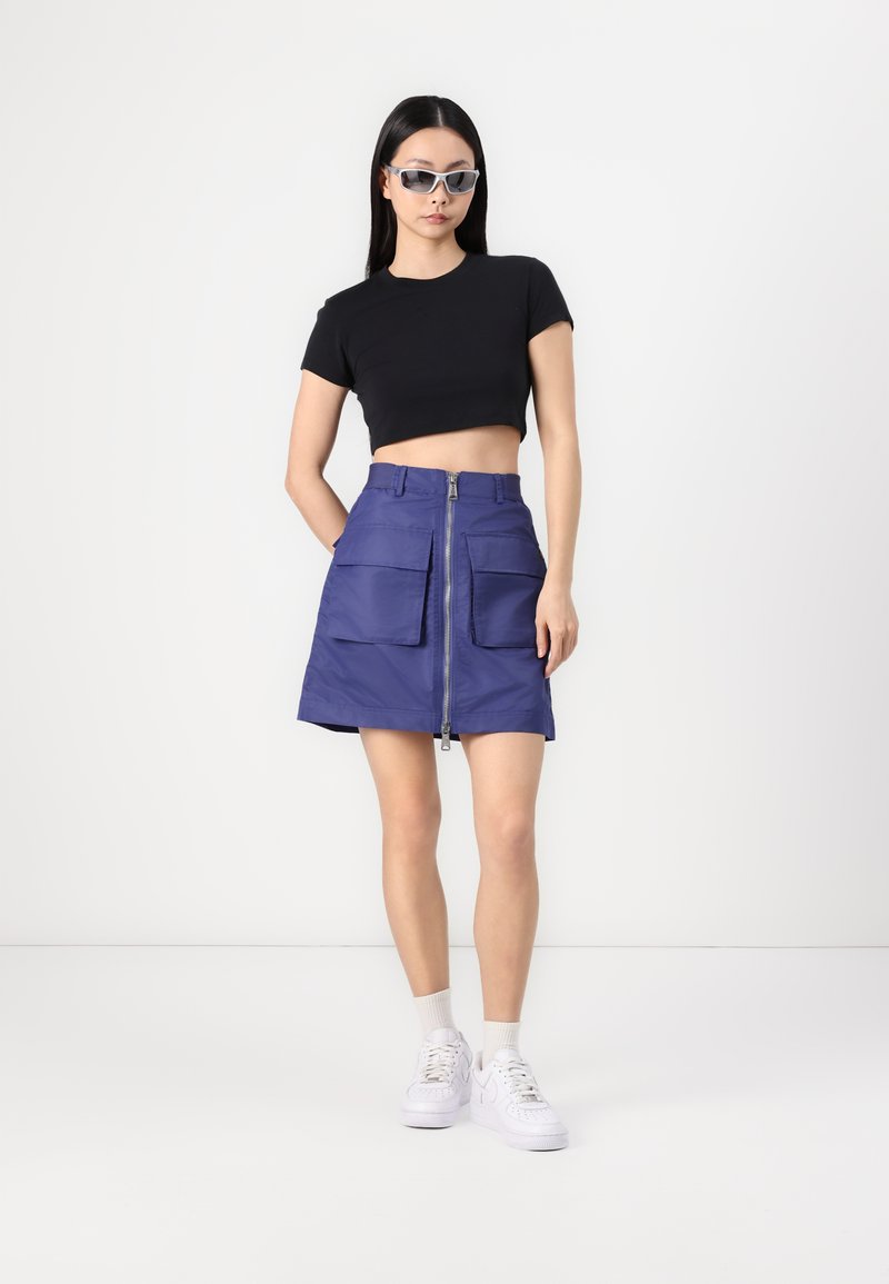 Black cropped shirt, purple skirt with two front pockets, zipper, and a smooth texture. White sneakers and clear sunglasses complete the look.