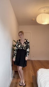 Juoda suknelė su ilgomis, pūstomis rankovėmis, dekoruotomis gėlių auksiniu raštu. Sijonas platėja, o iškirptė yra gili V formos.