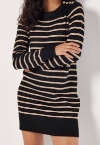 Femme portant une robe pull à manches longues rayée horizontalement en noir et beige avec des boutons dorés sur l'épaule, bras croisés.