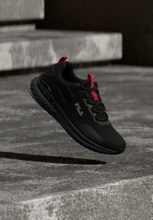 Črni tekaški čevelj Fila z rdečimi poudarki, oblazinjeno podplatom in logotipom na straneh, ki lebdi nad betonskimi stopnicami.