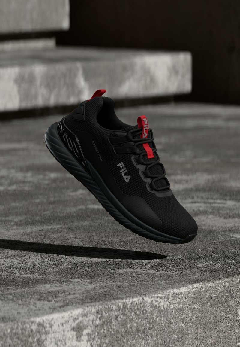 Chaussure de sport Fila noire avec des accents rouges, semelle rembourrée et côtés marqués survolant des marches en béton.