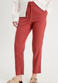 Pantalons moulants rouges en matière extensible avec une taille élastique à cordon, coupe slim et texture lisse. Associés à une chemise blanche à boutons et des sandales à glisser.