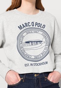 Grijze katoenen sweatshirt met een marineblauw logo-ontwerp en tekst, een ronde embleem en geribbelde halslijn, gedragen met donkere jeans.