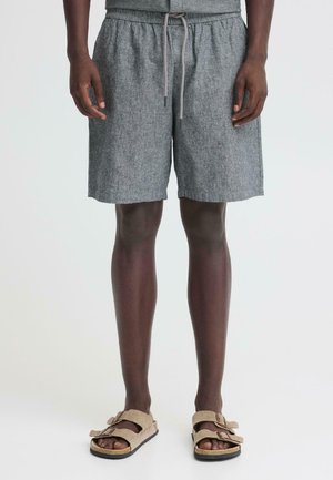 Mees, kes kannab halli nööriga lühikesi pükse ja beeže kahekordse rihmaga sandaale, seisab ühevärvse valge taustaga.