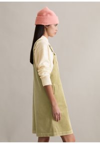 Sukienka typu ogrodniczki z sztruksu w jasnym zielonym kolorze, z regulowanymi ramiączkami, bocznymi kieszeniami, zestawiona z kremowym golfem i różową czapką beanie.
