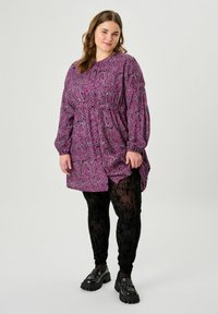 Abito viola con strisce zebra bianche e nere, maniche lunghe, vita arricciata, abbinato a leggings neri con motivi e mocassini neri spessi.