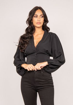 Blouse noire en tissu lisse, avec un col en V, une fermeture à boutons et des manches bouffantes. Portée avec un pantalon noir taille haute.