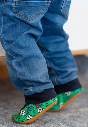 Blaue Jeans mit schmalem Schnitt und schwarzen Bündchen, kombiniert mit grünen, weichen Slippern, die mit fußballthemenähnlichen Mustern verziert sind.