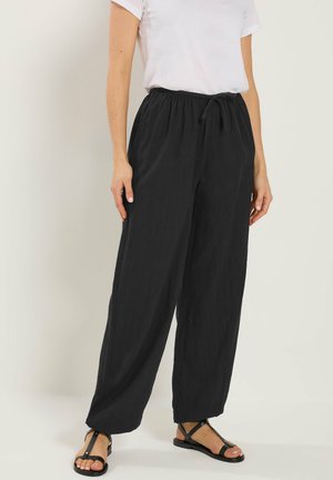 Kobieta ubrana w luźne czarne spodnie z troczkiem, białą koszulę z krótkim rękawem i czarne płaskie sandały, stojąca na tle jednolitego tła.