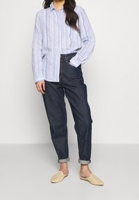 Camicia blu chiaro a righe bianche con bottoni, jeans in denim blu scuro e scarpe slip-on intrecciate beige, che mostrano una vestibilità rilassata e con i polsini arrotolati.