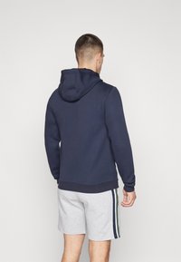 Felpa con cappuccio blu navy, con una superficie testurizzata, polsini e orlo a coste. Abbinata a pantaloni corti grigi chiari con strisce laterali verdi e blu navy.