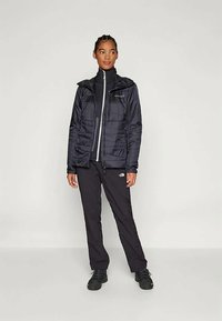 Chaqueta aislante negra con cierre de cremallera, capucha y un sutil patrón acolchado, combinada con pantalones negros ajustados y calzado negro.