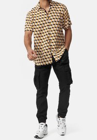 Kortärmad skjorta med gult och svart geometriskt mönster, kombinerad med svarta cargobyxor och vita sneakers. Knäpps med knappar framtill.