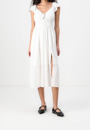 White maxi dress with short sleeves, gathered bodice, V-neck, and side slit. Paired with black shoes featuring decorative hardware.