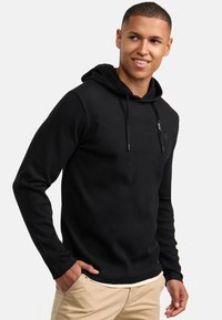 Sudadera con capucha negra de textura acanalada, con acentos de cordones y detalle de logo. Mangas largas y un ajuste relajado, combinada con pantalones beige.
