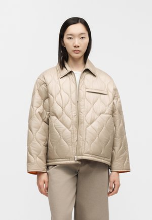 Jeune femme portant une veste matelassée beige zippée avec de grandes poches, debout devant un fond blanc uni.
