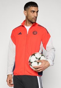 Αθλητικό μπουφάν σε κόκκινο και γκρι με φερμουάρ, λογότυπο FC Bayern και διαγώνιες ρίγες. Το μοντέλο κρατά μια μπάλα ποδοσφαίρου στο δεξί του χέρι.