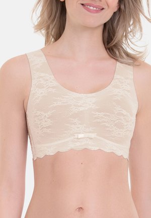 Woman wearing a beige lace bralette with a small bow in the center, smiling with loose hair over her shoulders.