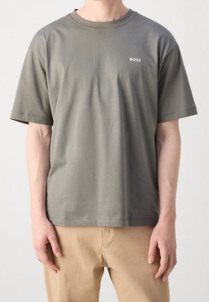 Personne portant un T-shirt à manches courtes vert olive avec le logo "BOSS" sur la poitrine et un pantalon beige, debout, les bras détendus le long du corps.