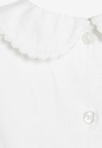 Weißes Kleidungsstück mit einem gewellten Kragen und zwei klaren Knöpfen. Der Stoff ist glatt, mit matter Oberfläche und ohne sichtbare Muster.