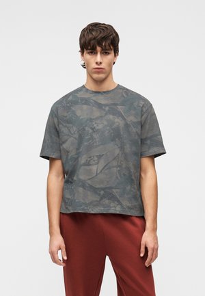Ung mand med kort brunt hår iført en løs grå T-shirt med camouflage mønster og røde bukser, stående mod en ensfarvet baggrund.