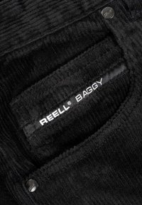 Black corduroy fabric with vertical ribbed texture, featuring a REELL® BAGGY label and a metal button accent near the pocket stitching.