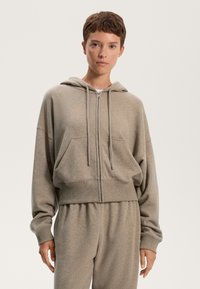 Felpa con zip accorciata in tessuto grigio chiaro con una texture morbida. Presenta un cappuccio con coulisse e tasche anteriori, abbinata a pantaloni coordinati.
