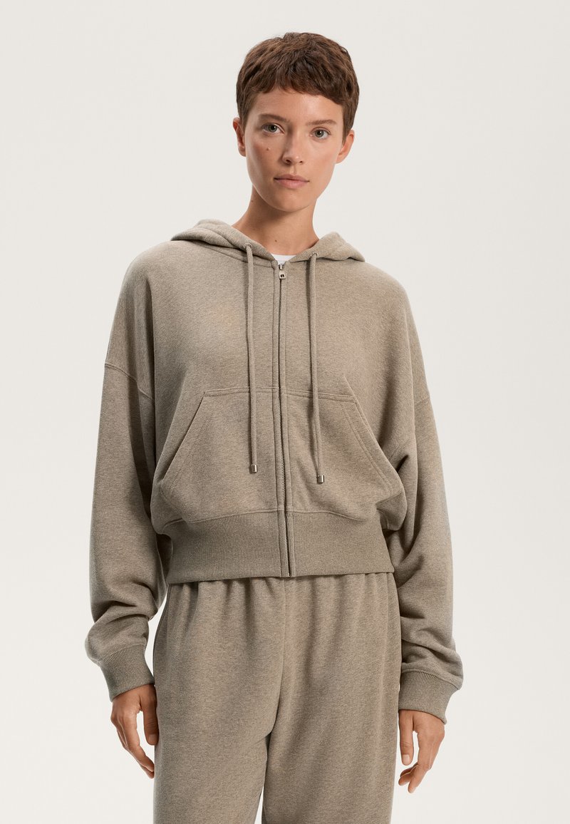 Felpa con zip accorciata in tessuto grigio chiaro con una texture morbida. Presenta un cappuccio con coulisse e tasche anteriori, abbinata a pantaloni coordinati.