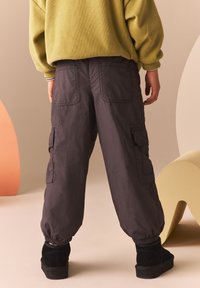 Pantalones cargo en gris oscuro con puños elásticos y múltiples bolsillos laterales, combinados con botas hasta el tobillo negras. Fondo en suaves colores pastel.