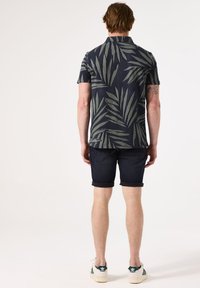 Kurzärmliges navyblaues Shirt mit grünem Blättermuster, kombiniert mit dunklen Denim-Shorts und weißen Sneakers mit grünen Akzenten.